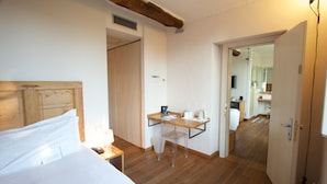 Habitación de hotel con cama de cabecera de madera, un escritorio con silla, una mesita con hervidor, y un baño visible a través de una puerta abierta.