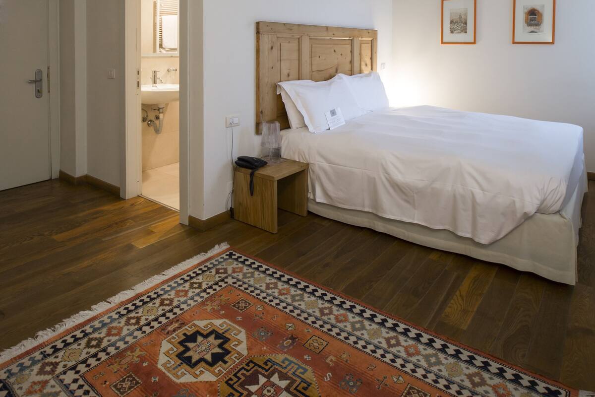 A bedroom with a wooden ceiling, a bed with white bedding, a nightstand, a rug, and a bathroom visible through an open door.
