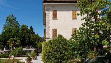 A building with a white facade and brown shutters, surrounded by lush greenery and potted plants.