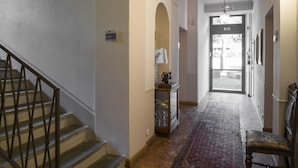 Un pasillo con una escalera, un candelabro, un banco y una puerta que da al exterior.