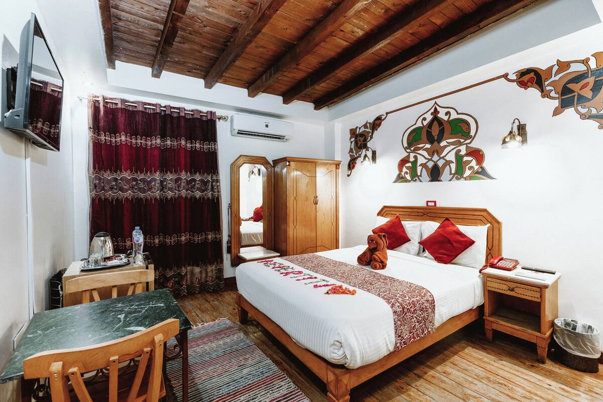 A bedroom with a wooden bed, a desk, and a chair. There is a mirror, a wall-mounted TV, and a decorative wall piece.
