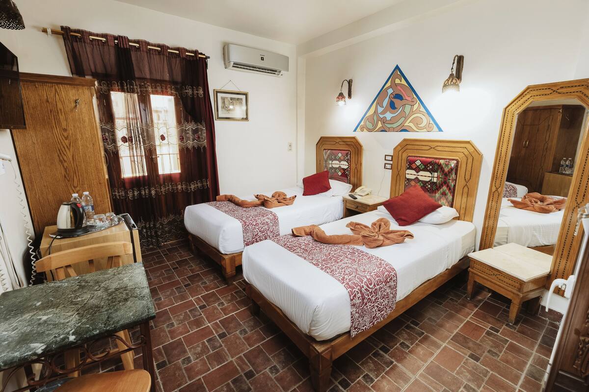 A hotel room with two beds, a small table, a bench, a mirror, and a wall-mounted air conditioner.