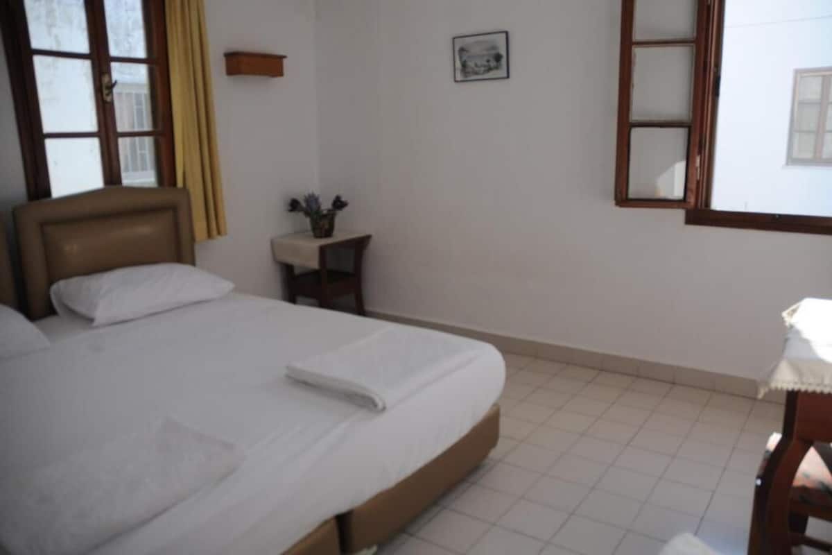 Une chambre à coucher avec un lit, une table de chevet avec une lampe, une fenêtre avec des rideaux et un tableau accroché au mur.