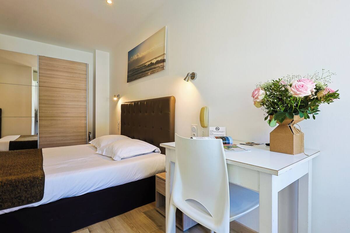 Une chambre d’hôtel avec un lit, un bureau, une chaise, un miroir, un tableau et un vase avec des fleurs.
