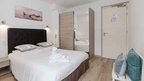 A modern hotel room with a large bed, a wooden headboard, a bedside table, a mirror, and a door with a bulletin board. - Cap Nègre Hôtel (Le Lavandou)