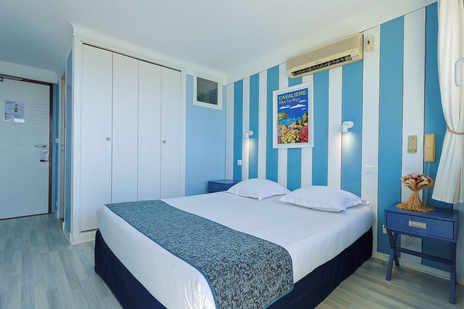 A hotel room with a bed, a nightstand, a mirror, a poster on the wall, and a door.