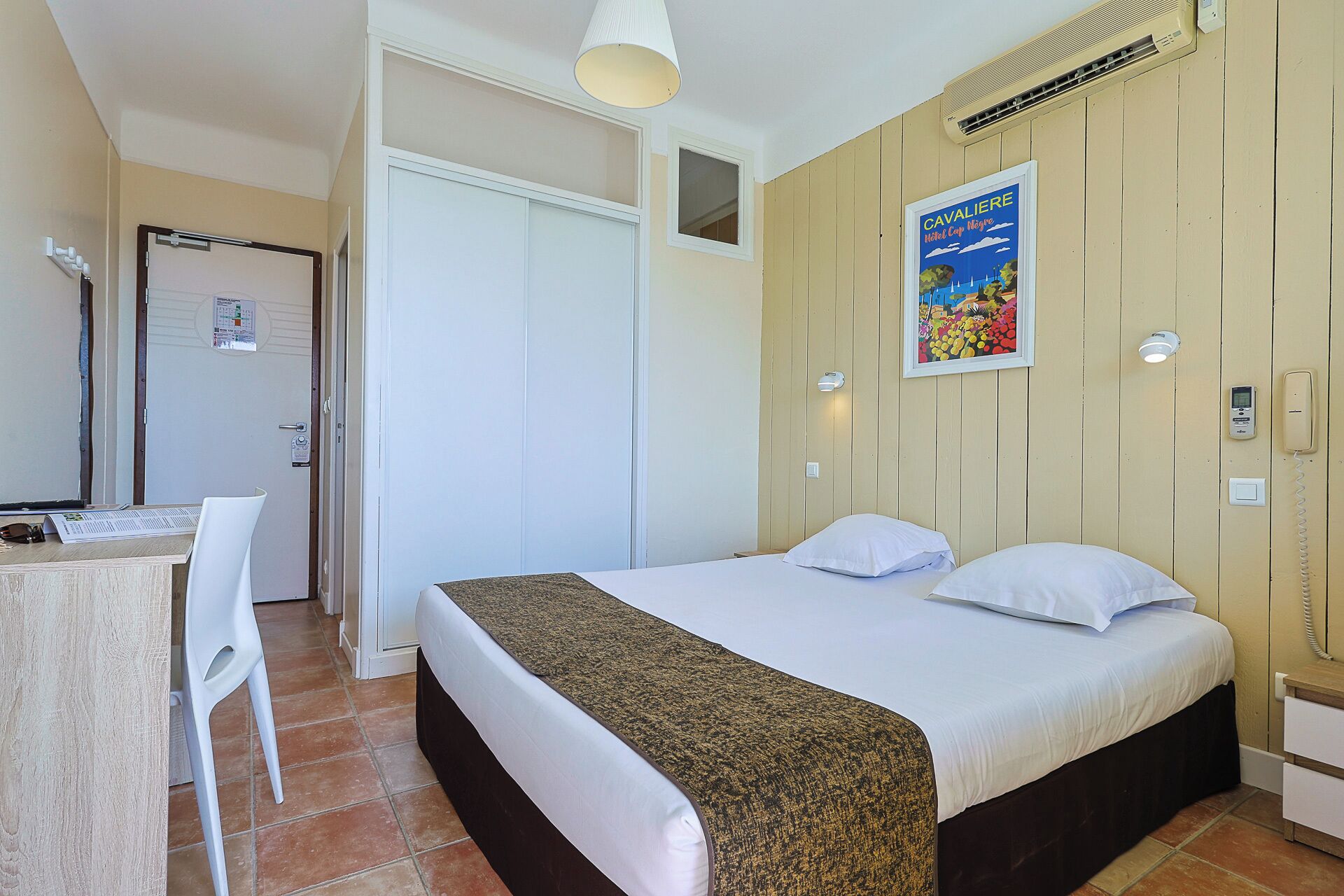 A bedroom with a bed, a desk, and a chair. There is a poster on the wall and a phone on the bedside table.