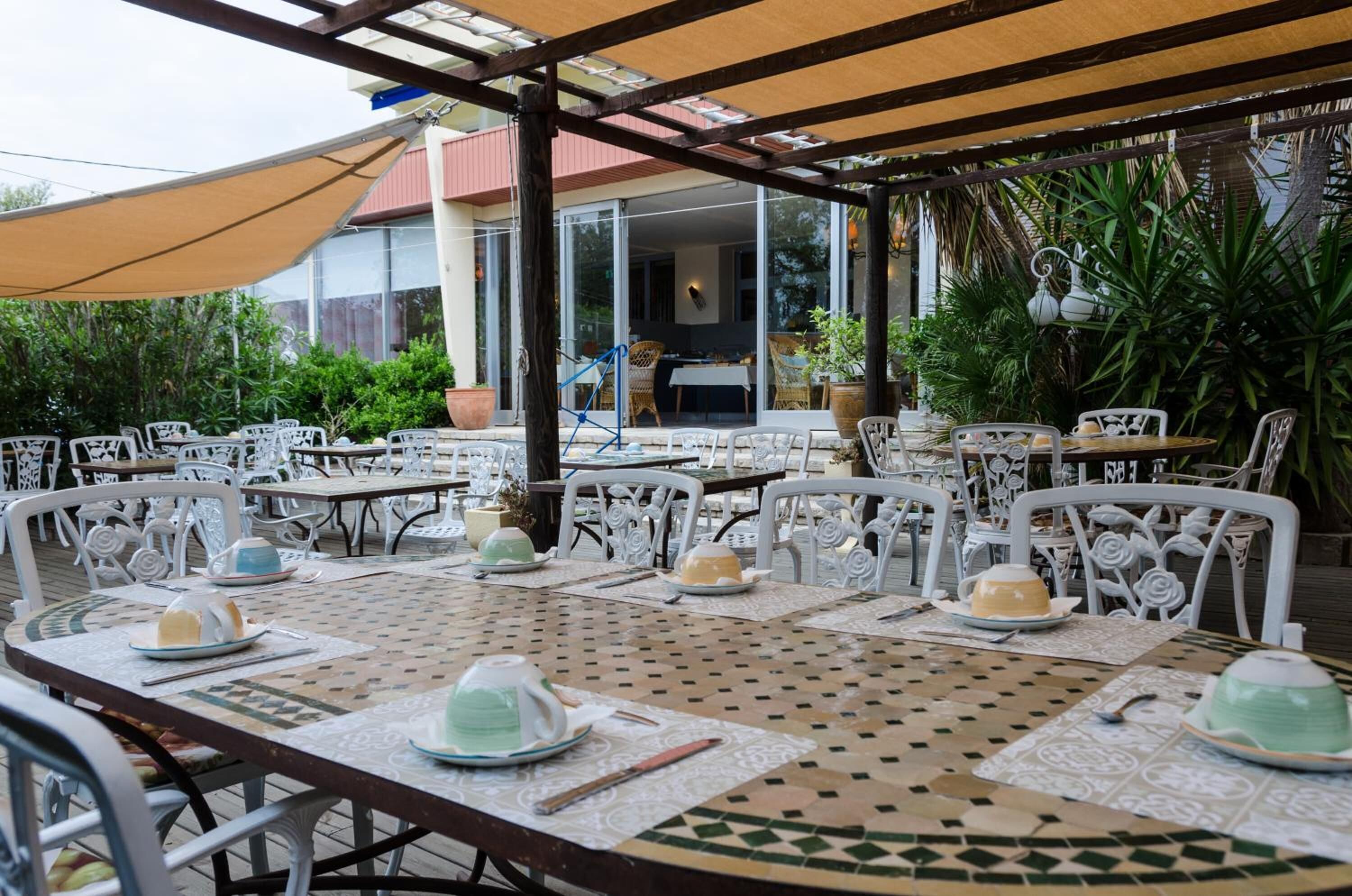 An outdoor dining area with white metal chairs and tables, covered by a pergola with a beige canopy.