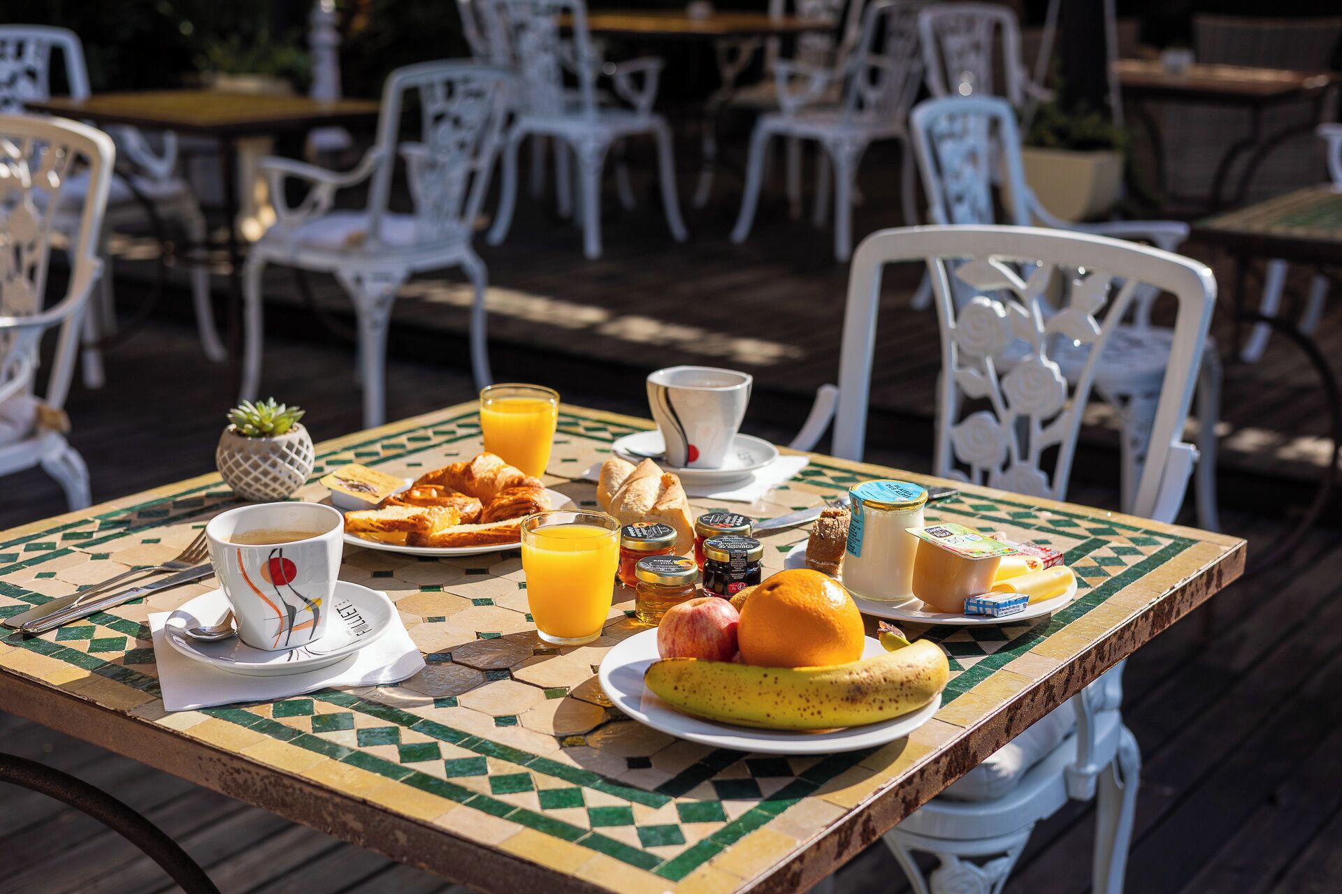 Una mesa dispuesta para el desayuno, con cruasanes, fruta y jugo, y sillas blancas al fondo.