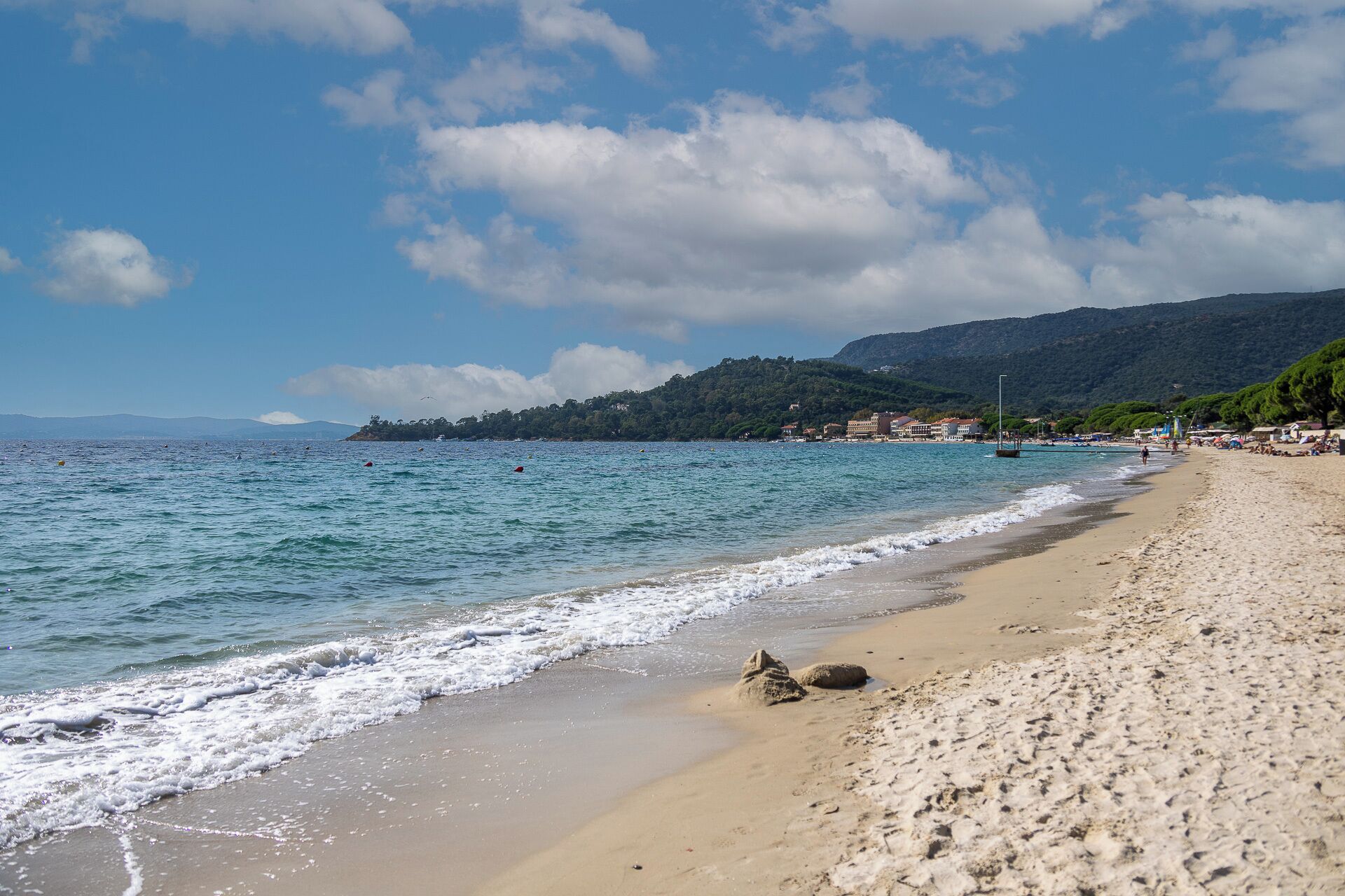 Una playa con agua azul cristalina, orilla de arena y una colina boscosa al fondo.