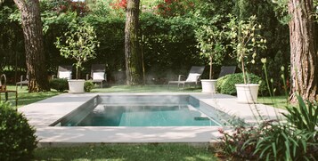 A rectangular pool with a hot tub in the center, surrounded by lush greenery and trees.