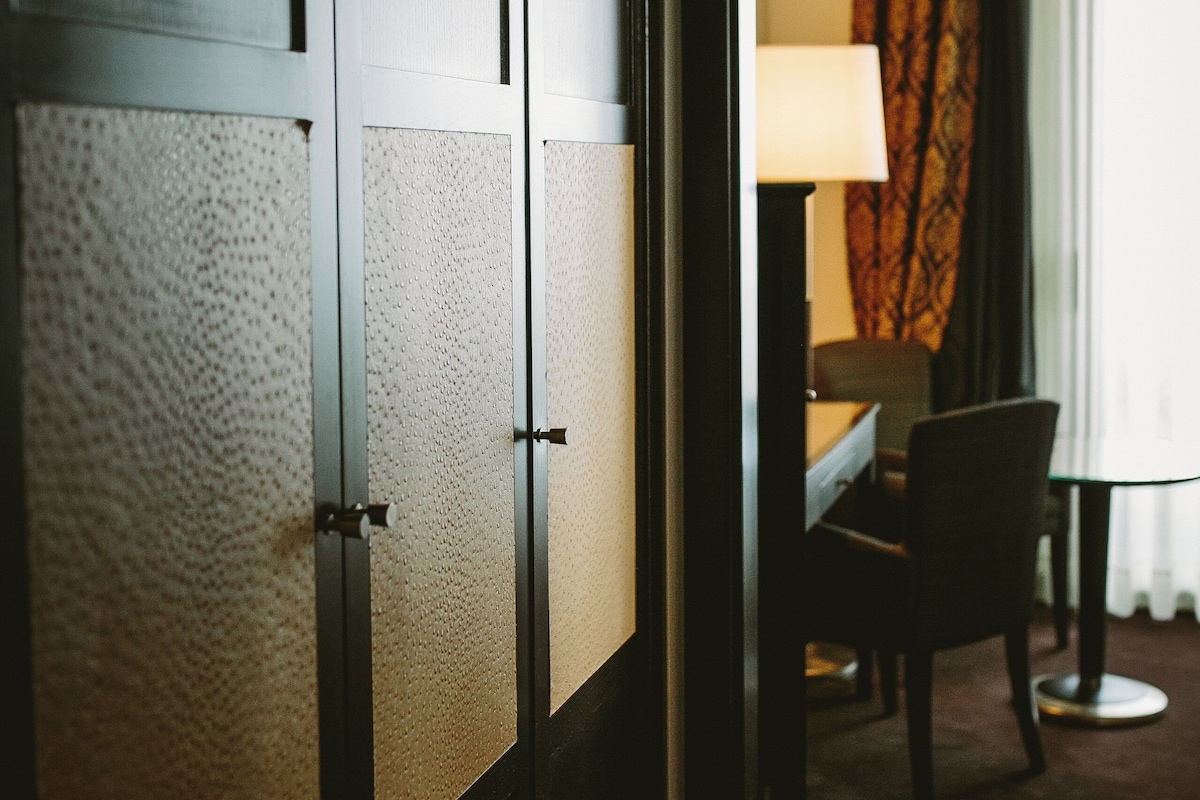 A hotel room with a dark wooden wardrobe, a desk with a lamp, and a chair.