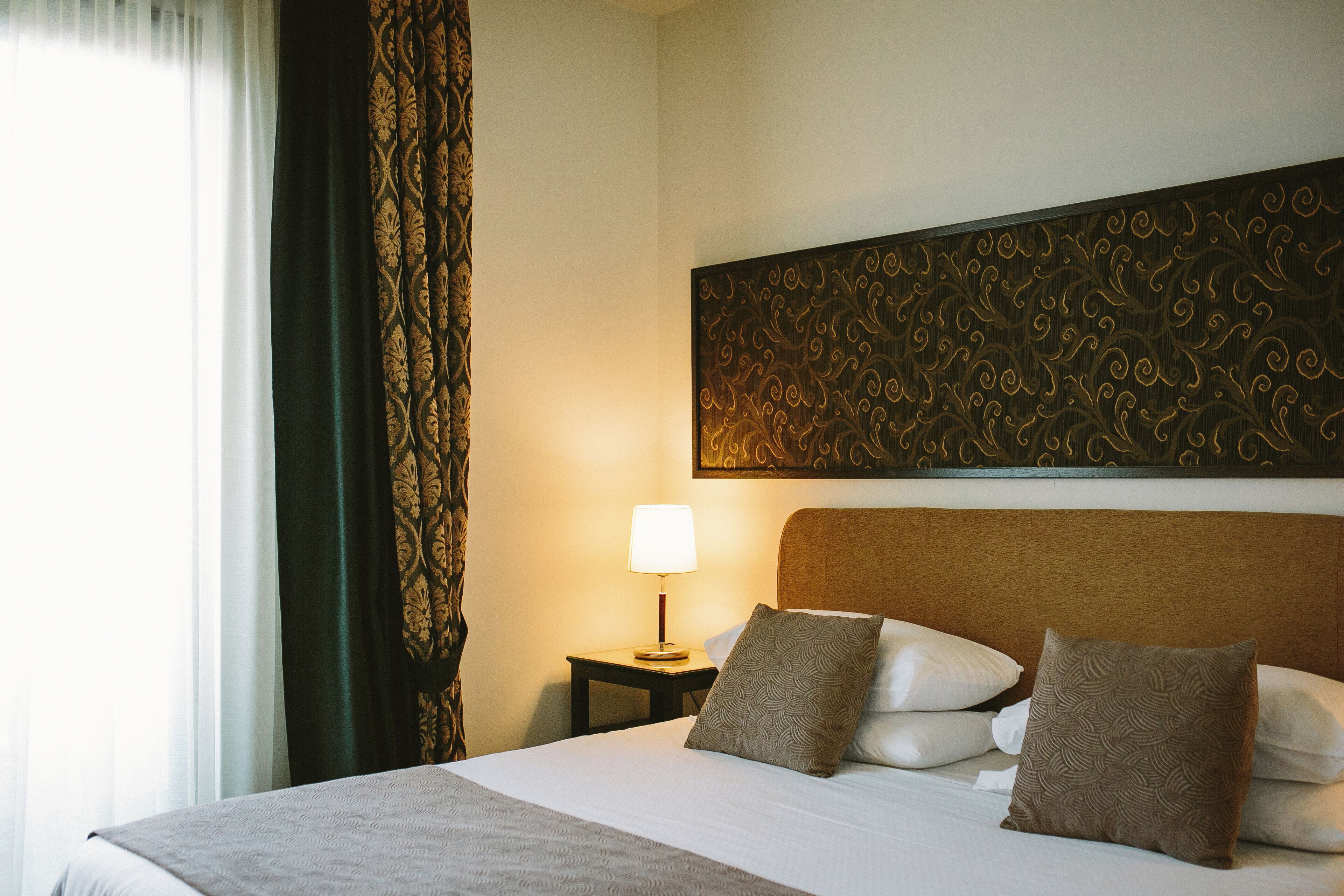Een hotelkamer met een groot bed, een nachtkastje met een lamp, kamergordijnen met een patroon, en een decoratief hoofdeinde.