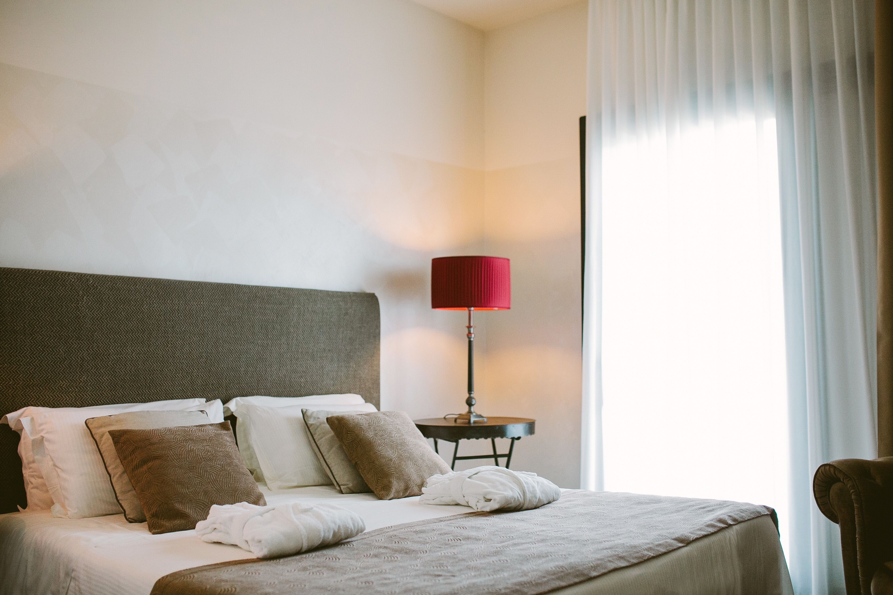 A neatly made bed with a grey headboard, a red lamp on a small table beside it, and a view of a window with sheer curtains.