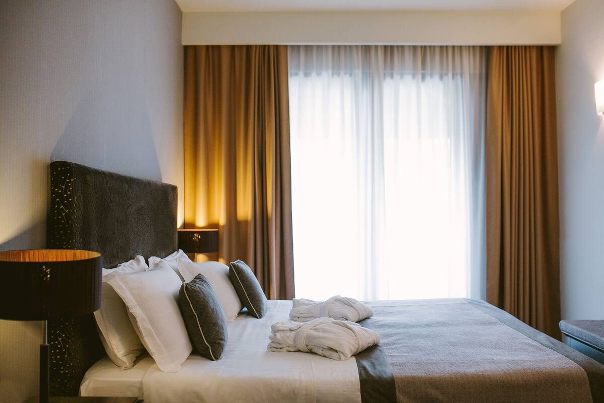 Ein ordentlich bezogenes Bett mit Kissen und Decke, eine Nachttischlampe und Vorhänge an einem Fenster.