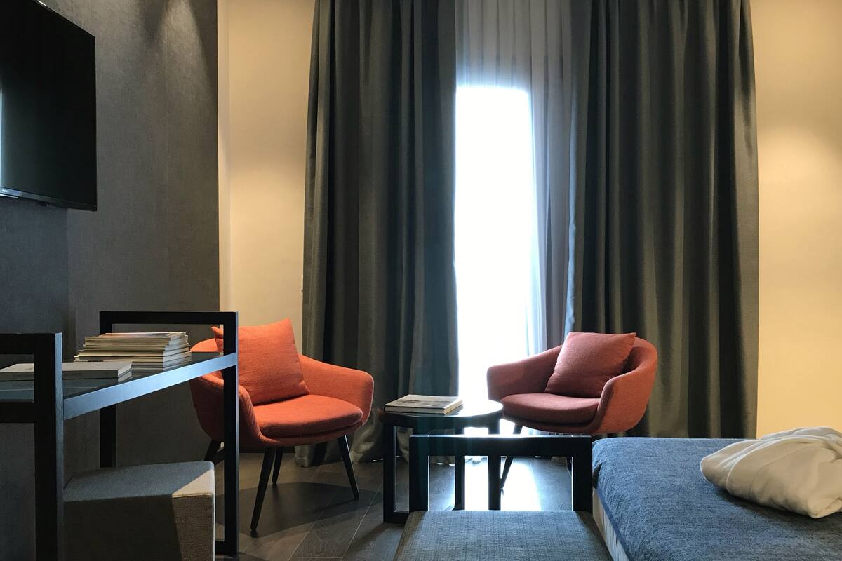 Ein modernes Hotelzimmer mit zwei roten Sesseln, einem kleinen Tisch, einer Couch, einem Fernseher und einem großen Fenster mit dunklen Vorhängen.