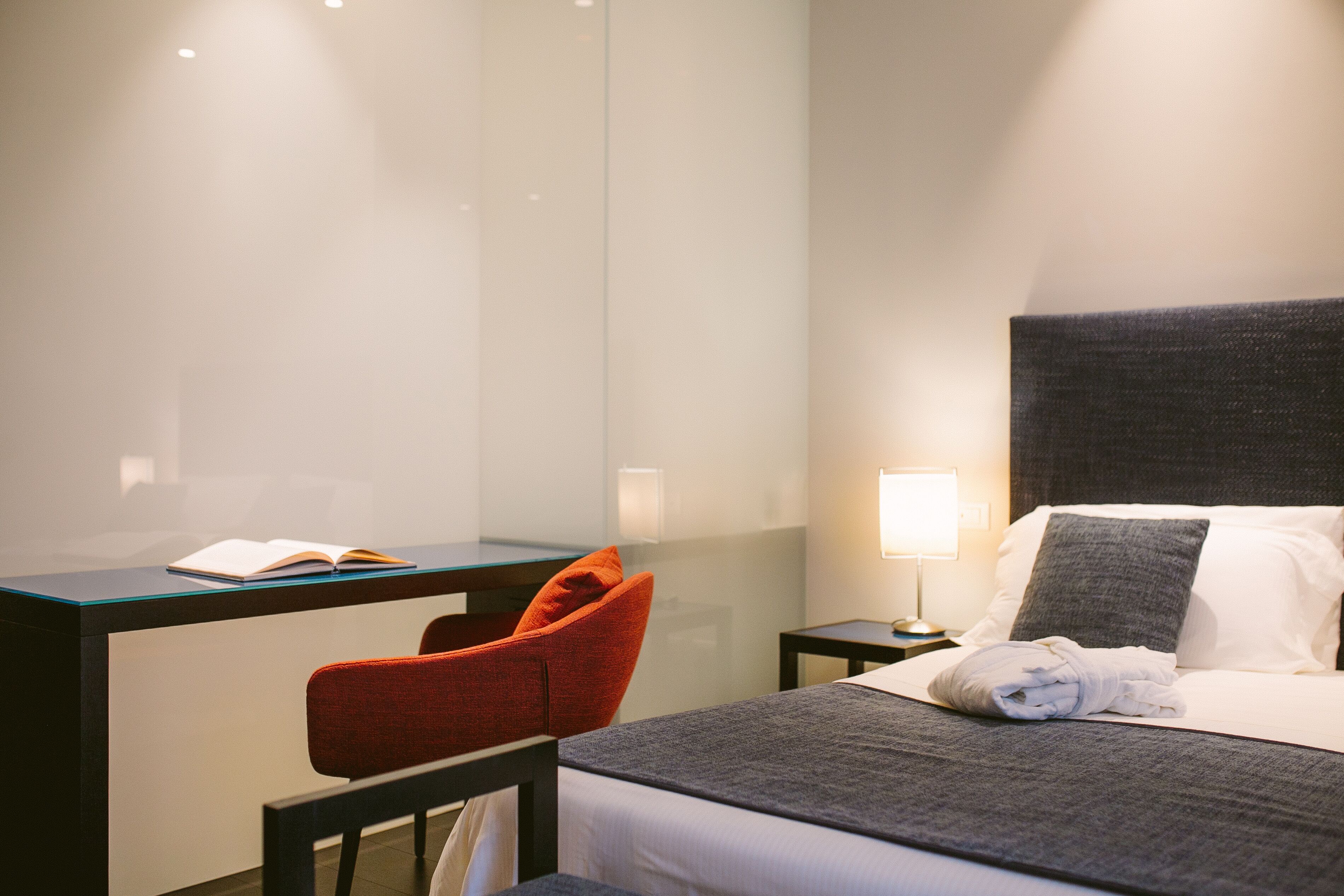 A hotel room with a bed, a desk, a chair, and a nightstand with a lamp.