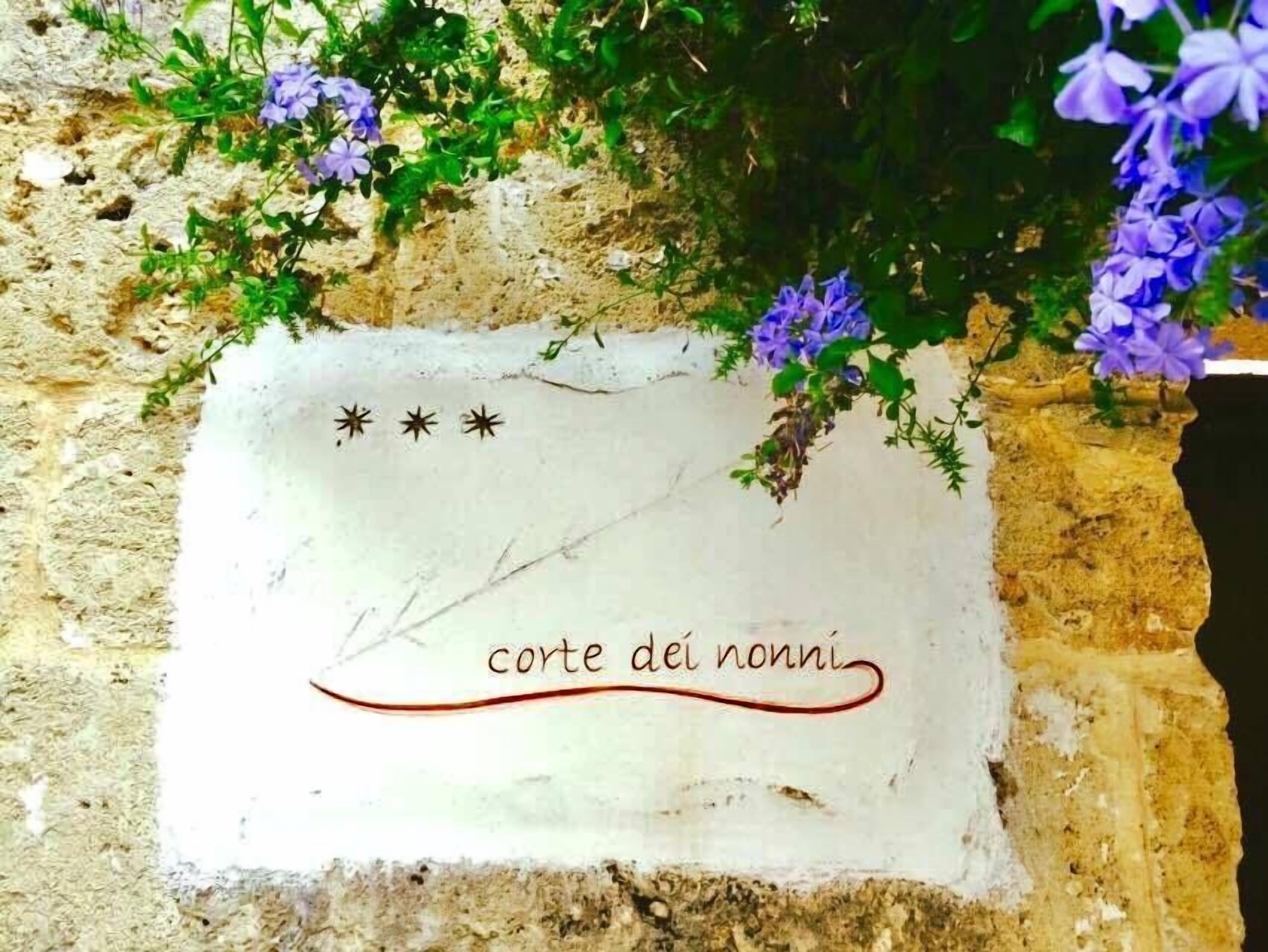 Eine Steinsäule mit einer weißen Plakette, auf der "corte dei nonni" steht, umgeben von violetten Blüten.