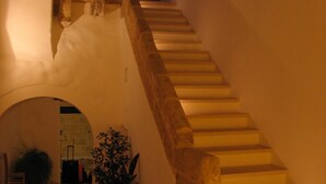 Uma escadaria com balaústres de pedra que leva a um andar superior, cercada por paredes brancas e um pequeno arco.