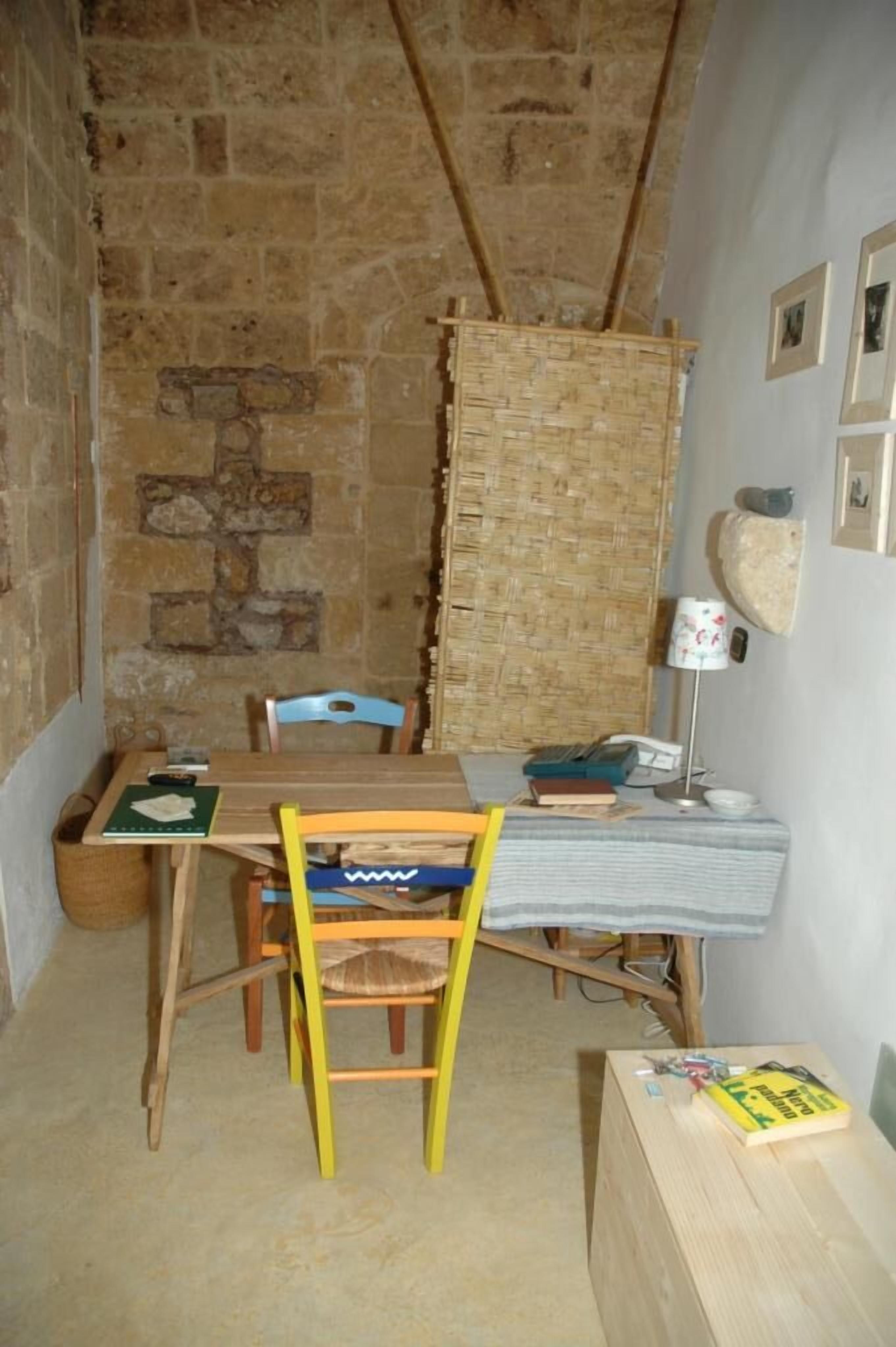 A small, rustic room with a stone wall, a wooden table, a chair, and a bed with a striped blanket.
