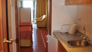 A compact kitchen with a sink, toaster, and refrigerator. A hallway leads to a bedroom with a bed and a chair. - Vila Marta (Funchal)