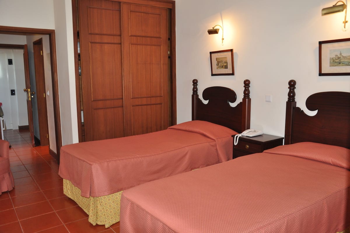A room with two wooden beds, a wooden wardrobe, a bedside table with a phone, and a wall-mounted light fixture.