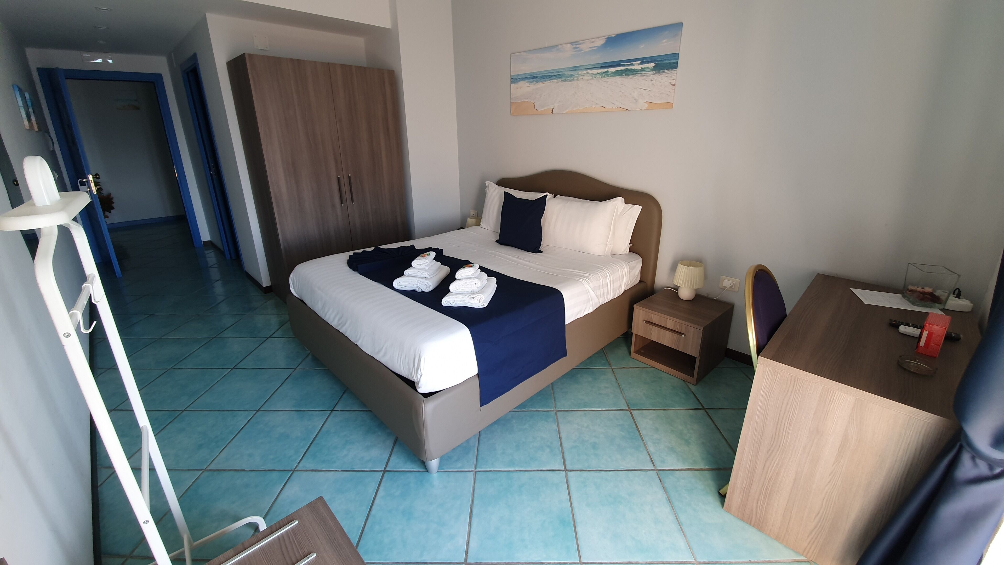 Ein Hotelzimmer mit Bett, Nachttisch, Schreibtisch, Kleiderschrank und einem Bild mit Strandmotiv.
