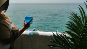 Eine Person hält ein blaues Getränk und blickt auf das ruhige Meer.