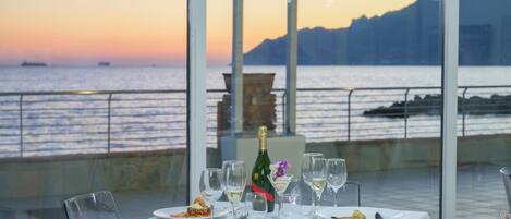 Una mesa dispuesta para una comida, con una botella de champán, copas y platos de comida, con vistas a un cuerpo de agua y una cadena montañosa distante.
