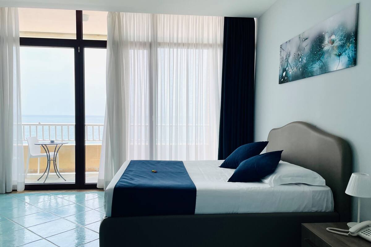 Habitación de hotel con una cama, una mesita de noche con lámpara y vista al mar a través de amplias ventanas.