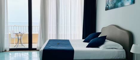 A hotel room with a bed, a bedside table with a lamp, and a view of the sea through large windows.