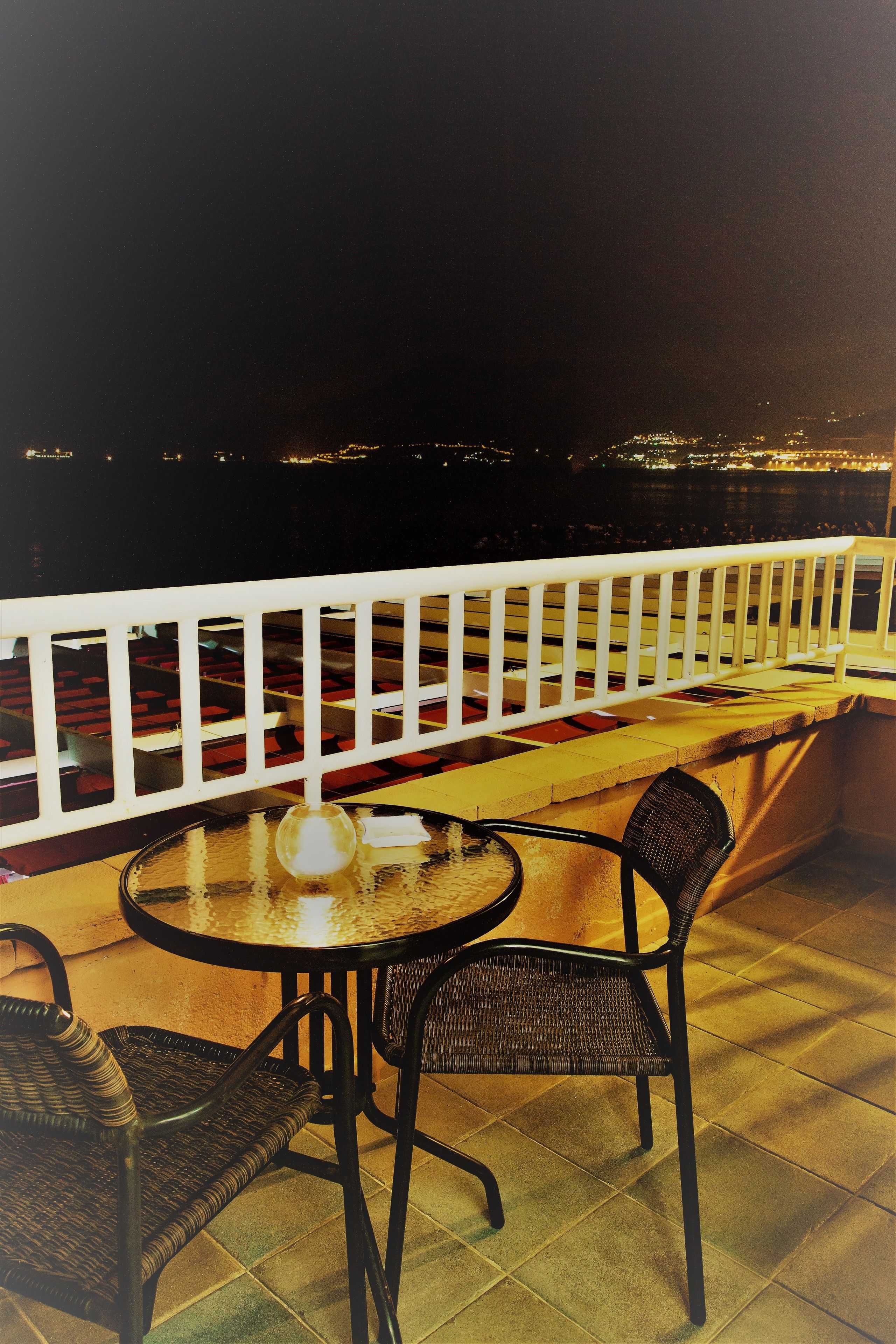 A balcony with a round table and two chairs, a white railing, and a view of a cityscape at night.