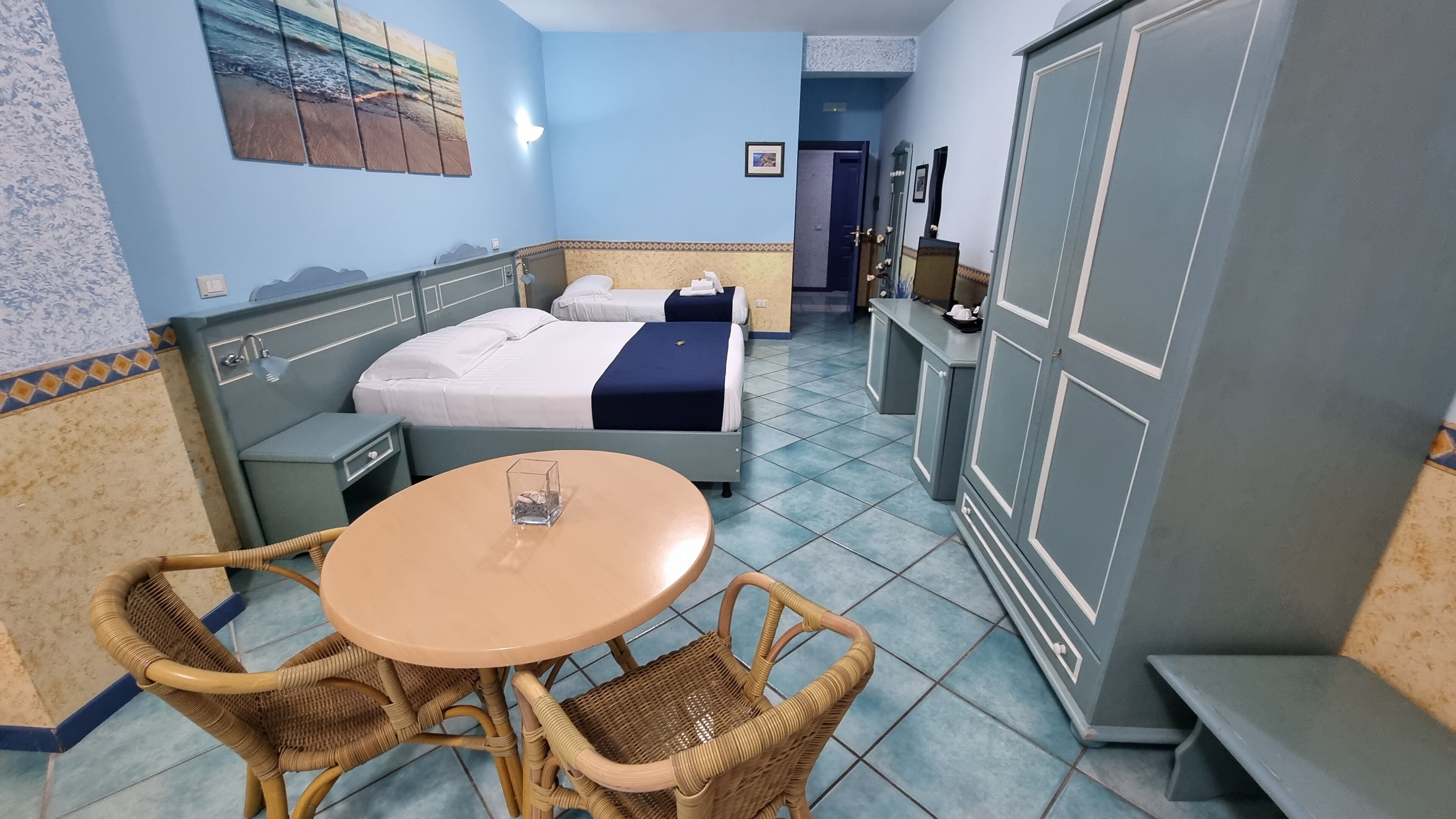 A hotel room with two beds, a small table, wicker chairs, a bathroom with a mirror, and a wardrobe.