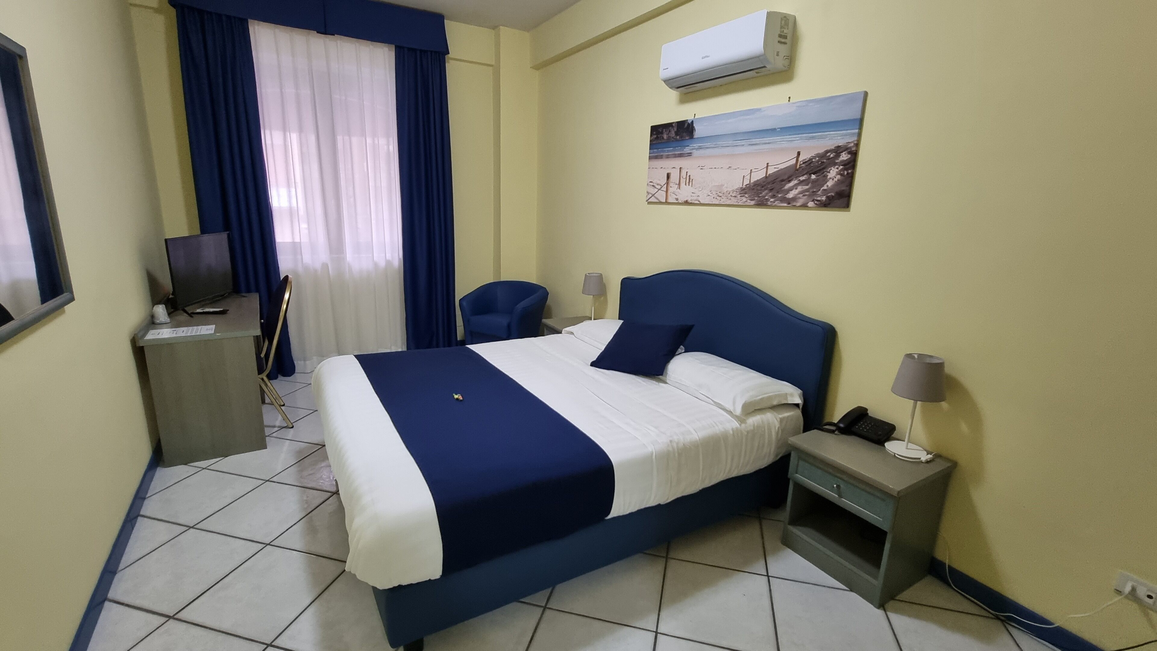 A hotel room with a bed, a desk, a chair, a TV, an air conditioner, and a painting on the wall.