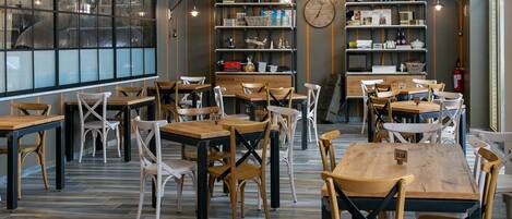 A modern cafe with wooden tables and chairs, shelves with books and bottles, and a large window.