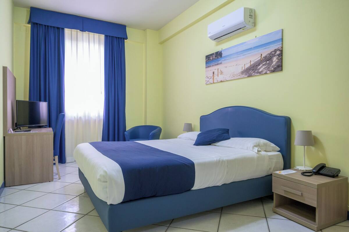 Habitación de hotel con cama, escritorio con televisor, silla, mesita de noche con teléfono y equipo de aire acondicionado.