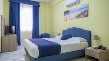 A hotel room with a bed, a desk with a TV, a chair, a nightstand with a phone, and an air conditioning unit.