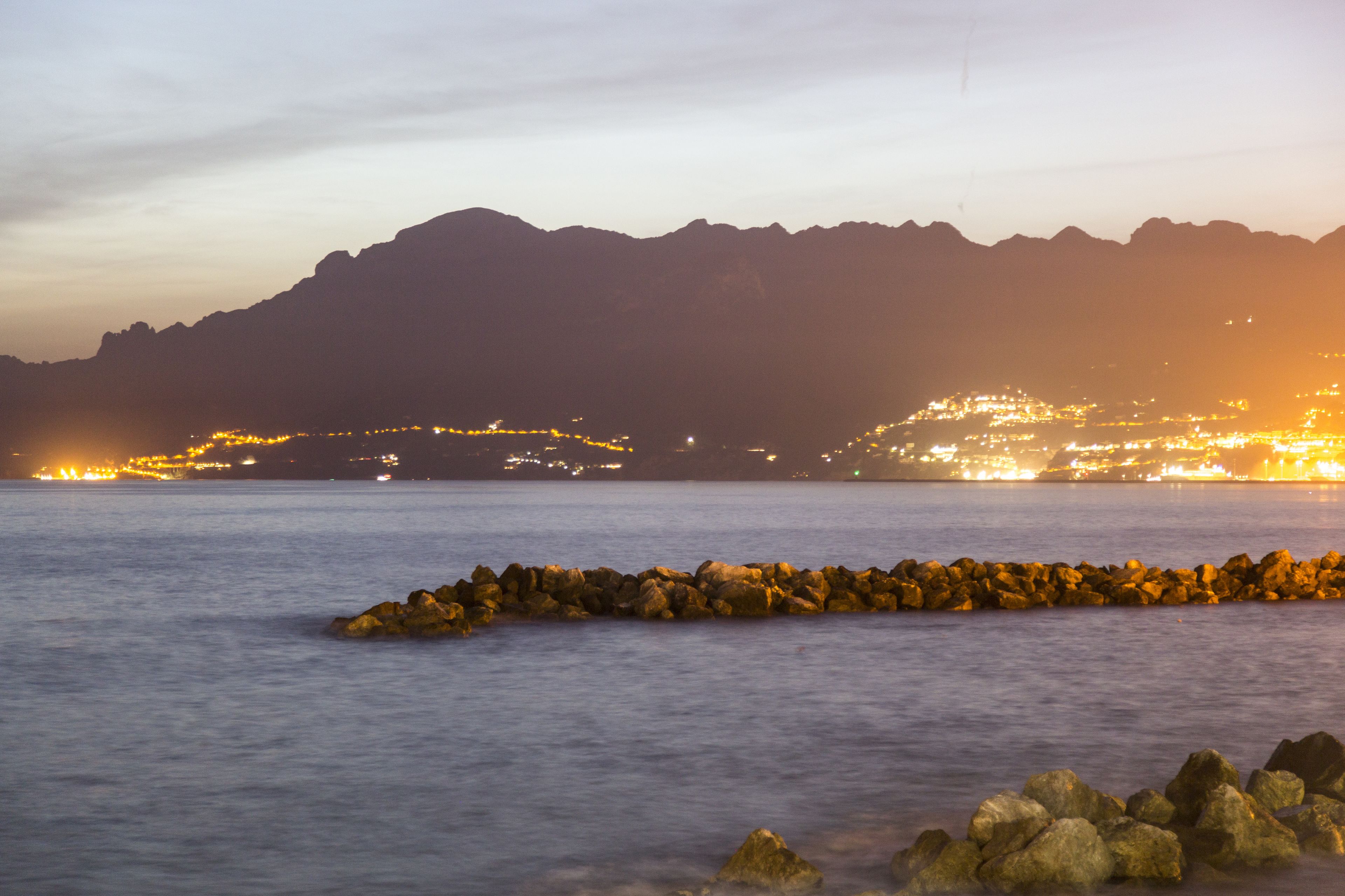 A coastal landscape with a rocky breakwater in the foreground and a city illuminated in the background.