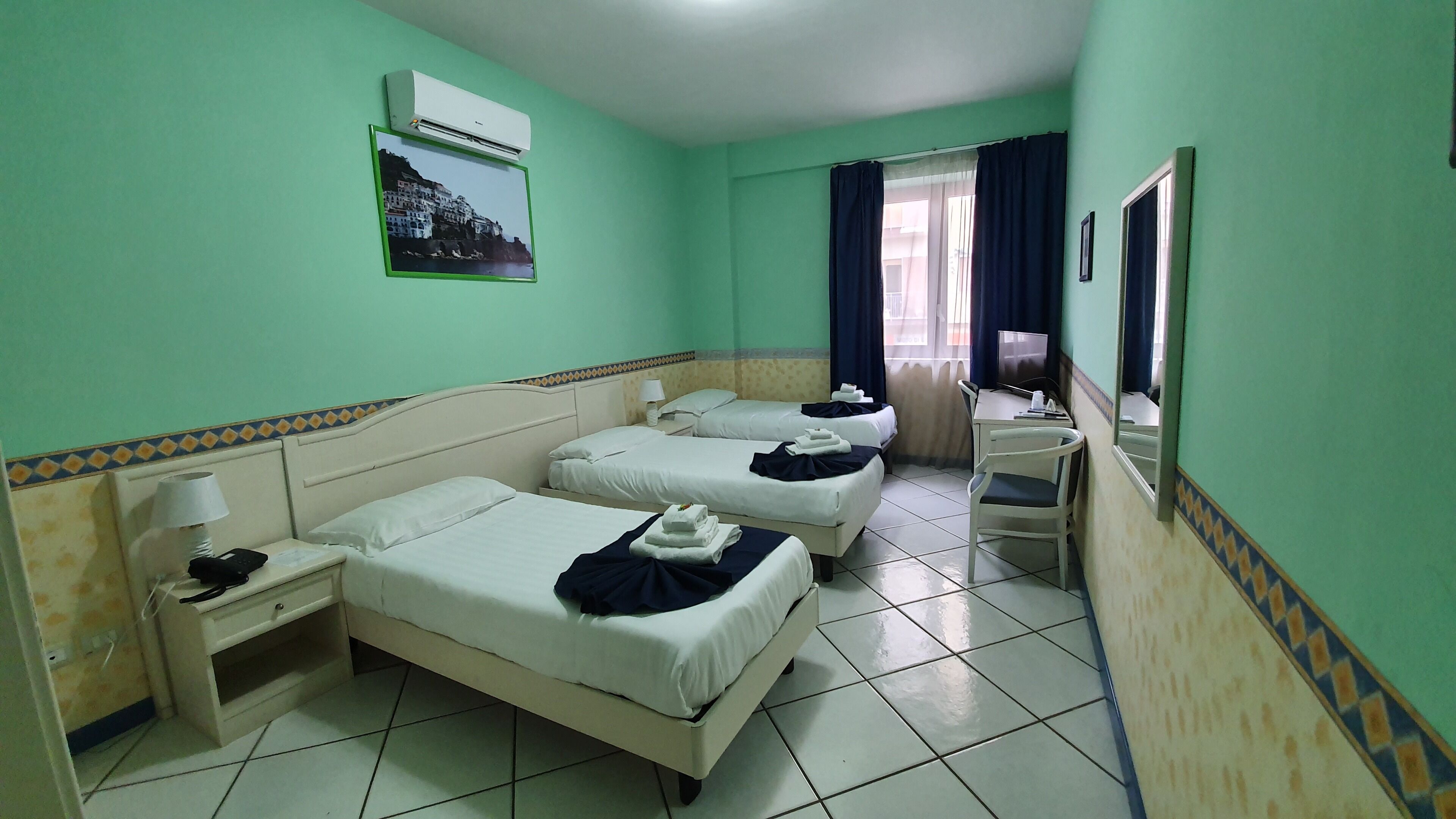 Habitación de hotel con tres camas, un escritorio, una silla, aire acondicionado y una ventana con cortinas.