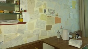 Un mur encombré, couvert de nombreuses notes et illustrations manuscrites, un comptoir en bois avec une machine à café, et une étagère ornée d’objets décoratifs.