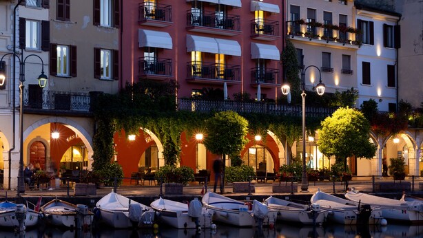 Un hotel frente al agua con un letrero que dice 'HOTEL PIROSCAPO'. Hay barcos amarrados en el agua frente al hotel.