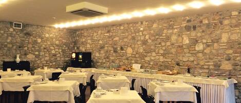 Un salón de banquetes con paredes de piedra, varias mesas dispuestas con manteles blancos y una pantalla de televisión al fondo.