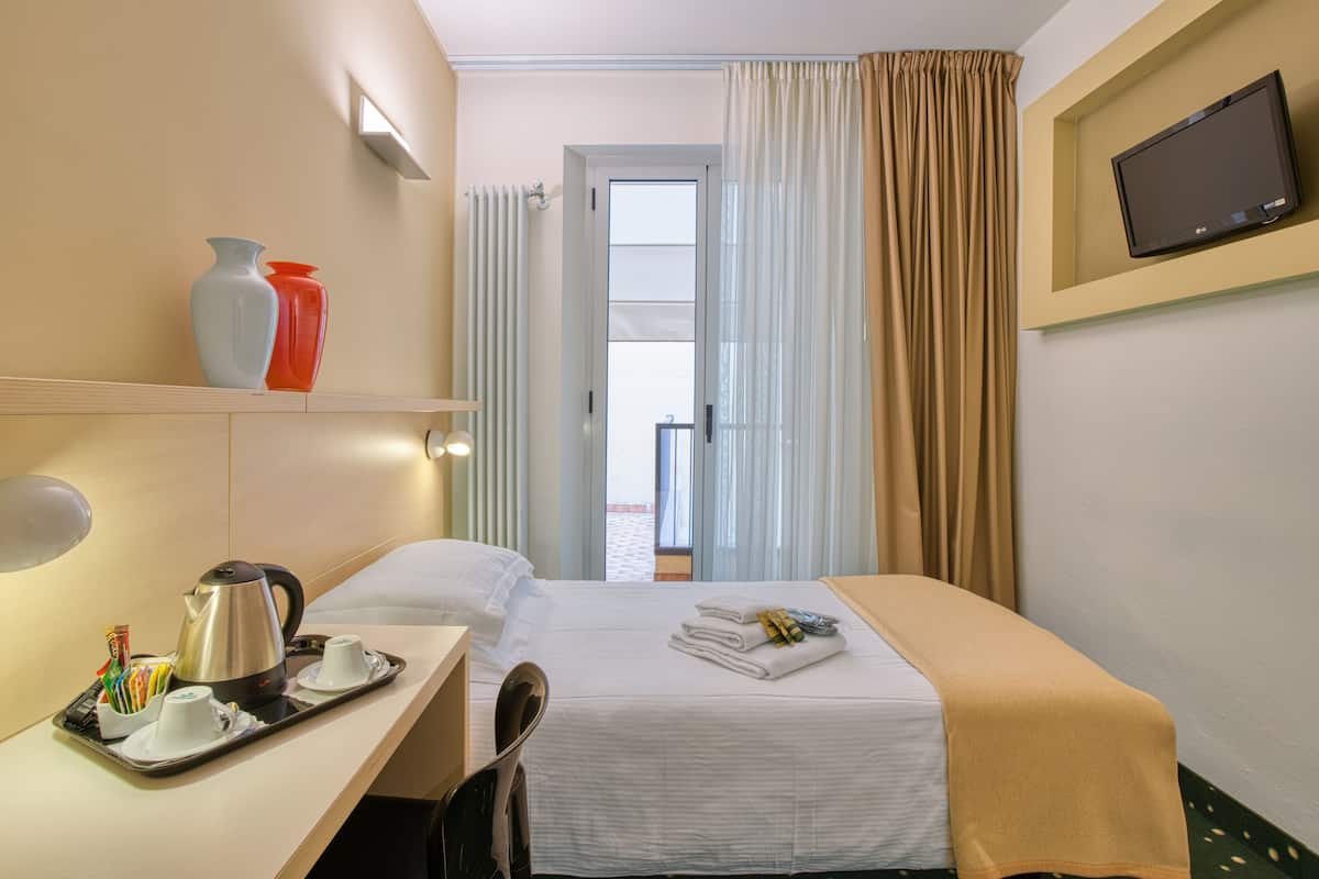 Habitación de hotel con cama, escritorio con tetera y tazas, televisor en la pared y vista al exterior por la ventana.