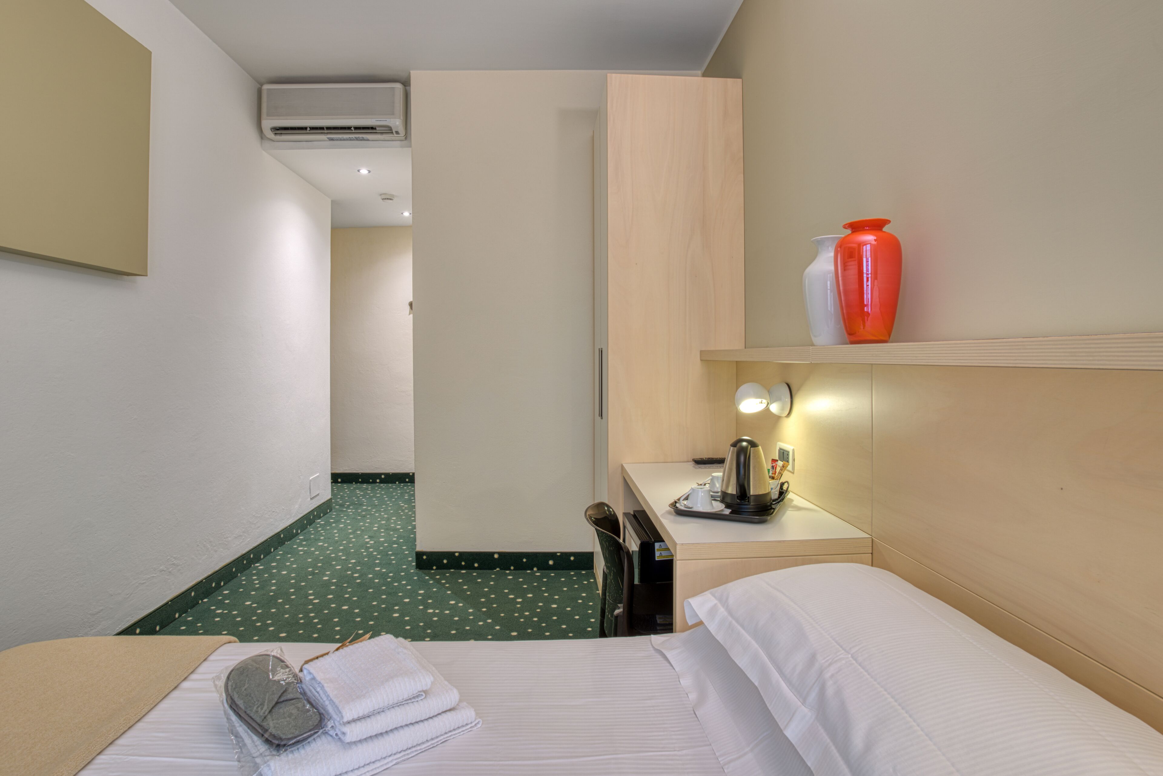 A hotel room with a bed, a desk with a lamp, a phone, and a vase on the nightstand.