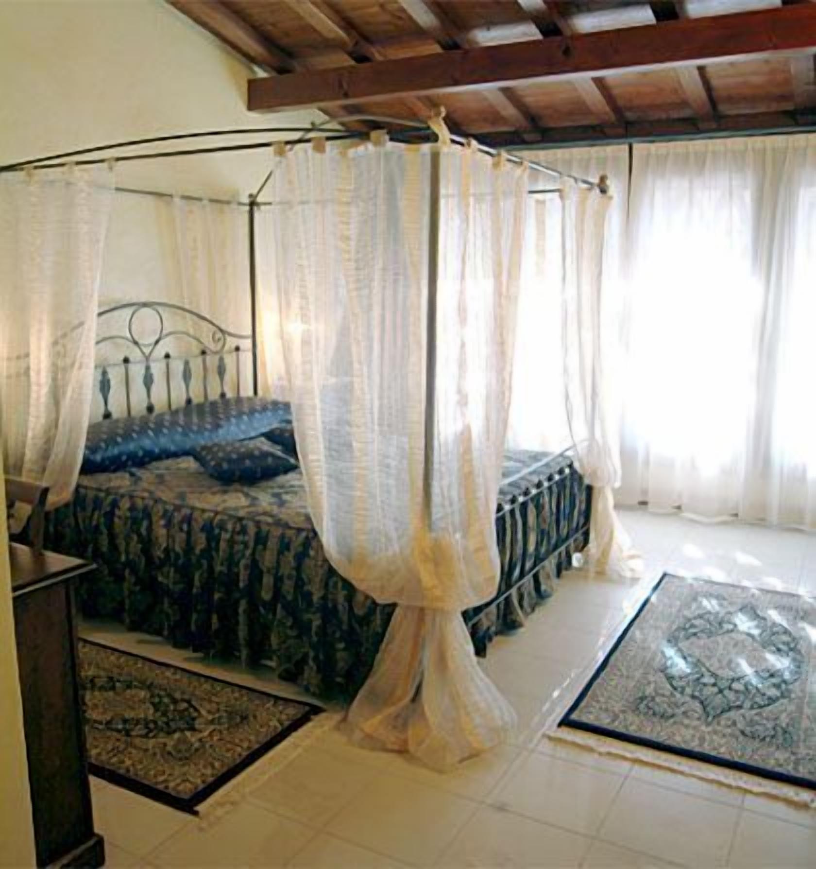 A bedroom with a four-poster bed, a wooden ceiling, and a large window with sheer curtains.