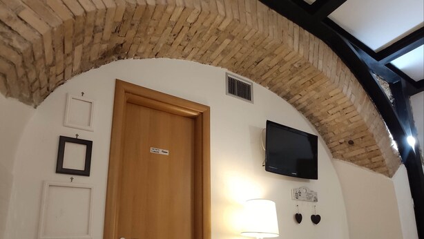 Una habitación con puerta de madera que indica "VETTOI FUNARDO", un televisor montado en la pared, una mesa con cestas y una colcha estampada.