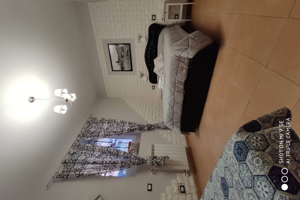 A bedroom with a bed, a nightstand, a wall-mounted lamp, and a patterned curtain.