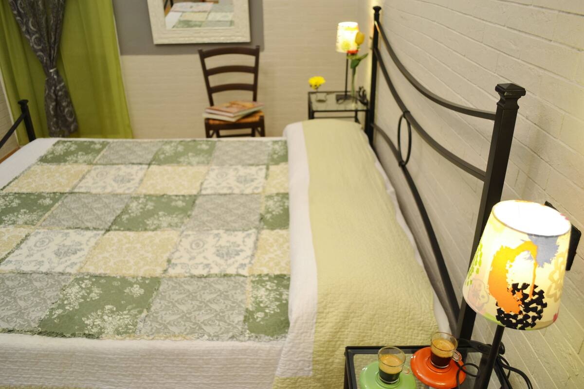 A neatly made bed with a quilt, a bedside table with a lamp, and a chair with a book.