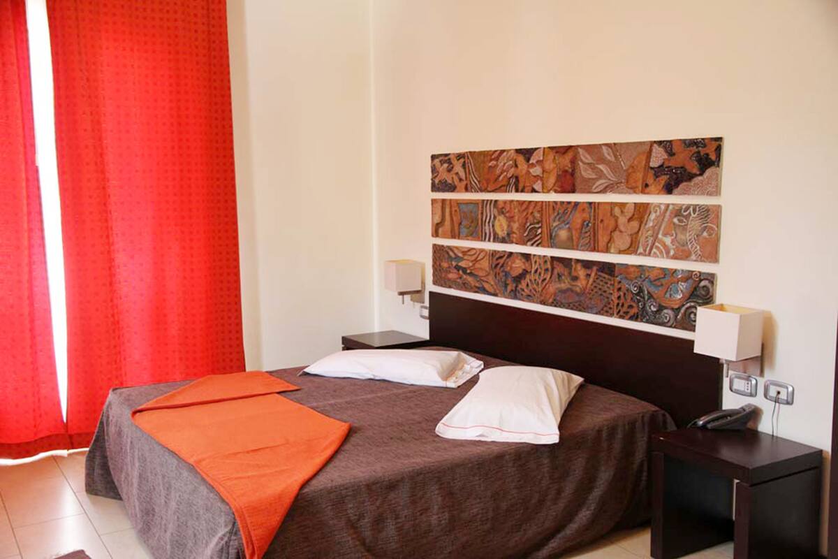 A hotel room with a large bed, two bedside tables, a wall-mounted artwork, and a red curtain.