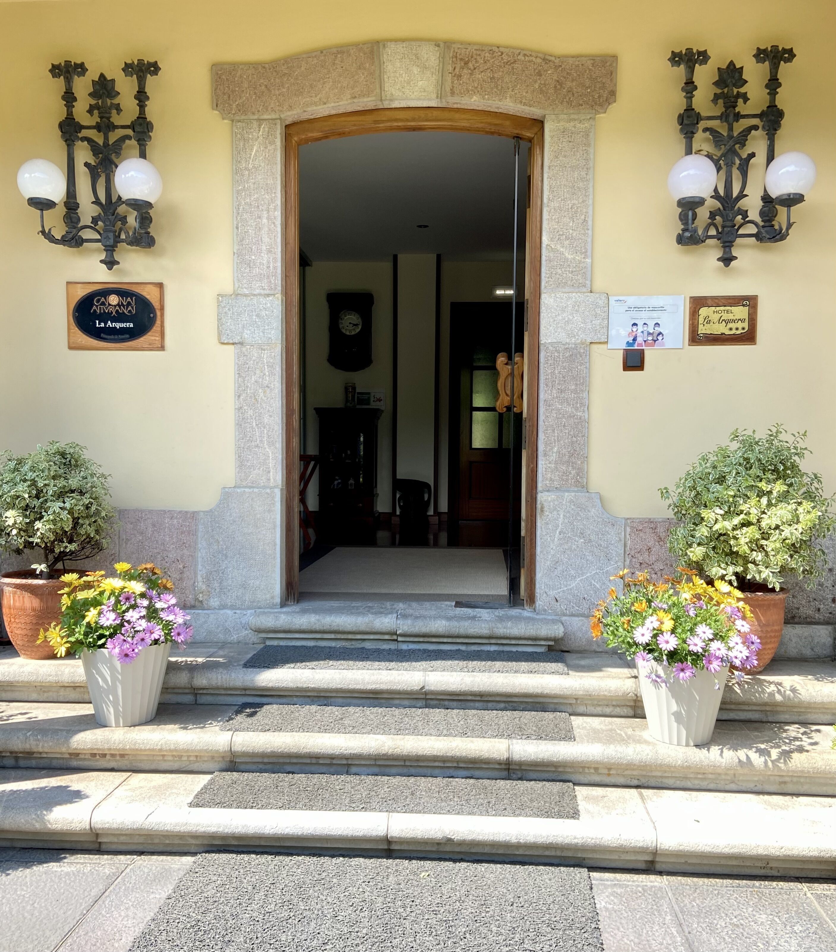 Entrada de hotel con un arco de piedra, un reloj y dos macetas con flores.